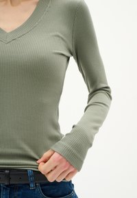 Olivgrön ribbad långärmad topp med V-ringning. Har en figurnära design och strukturerad tyg. Kombineras med ett svart bälte och blå jeans.