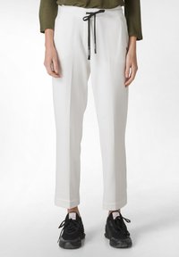 Pantalones blancos de corte relajado, con una cintura ajustable de cordón negro, bolsillos laterales y bajos con puños. Llevados con zapatillas negras.