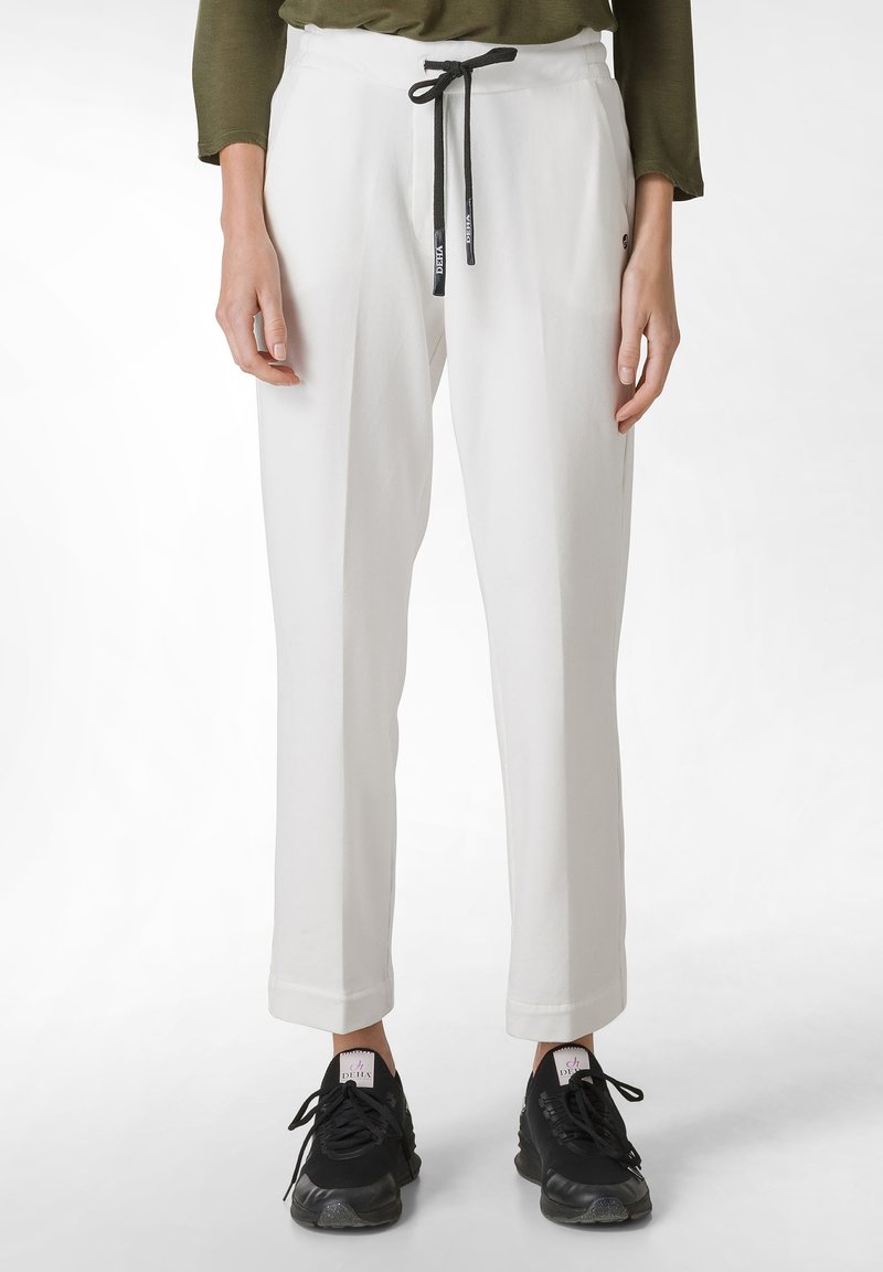 Pantalones blancos de corte relajado, con una cintura ajustable de cordón negro, bolsillos laterales y bajos con puños. Llevados con zapatillas negras.