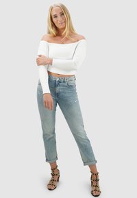 Witte off-the-shoulder blouse met lange mouwen, lichtblauwe distressed jeans met opgerolde boorden, zwarte hoge hakken met gouden studs.