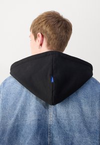 Capucha negra en una chaqueta de denim, con un diseño puntiagudo, texturas contrastantes y una pequeña etiqueta de logo azul en el lado izquierdo de la capucha.