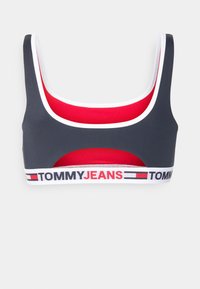 Sutien desportivo com um corpo cinza, forro vermelho, acabamento branco e uma faixa elástica com a marca que diz "TOMMY JEANS" em cores contrastantes.