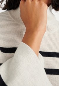 Svetle šedý, žebrovaný pletený svetr s námořnickými horizontálními pruhy. Volné rukávy. Jemná textura. Detailní záběr ruky položené poblíž límce.