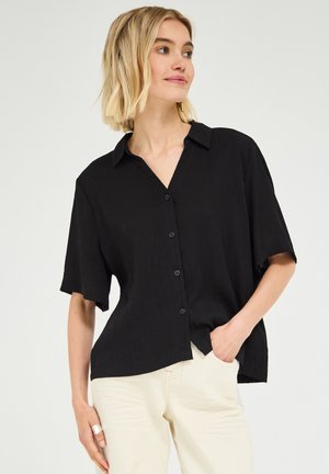 Femme aux cheveux blonds courts portant une chemise noire ample à boutons et un pantalon clair, main dans la poche, regardant légèrement vers la droite.