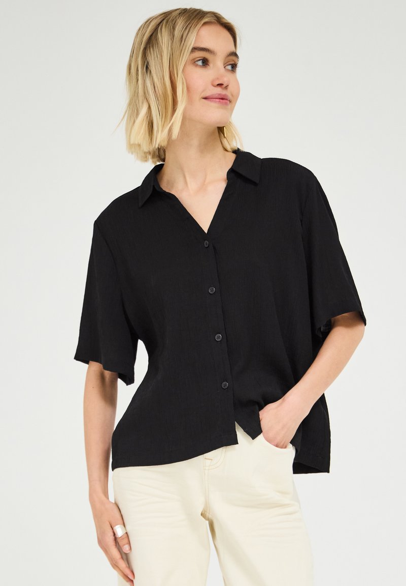 Femme aux cheveux blonds courts portant une chemise noire ample à boutons et un pantalon clair, main dans la poche, regardant légèrement vers la droite.