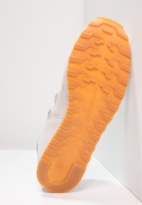 Semelle de chaussure de sport grise avec une base en caoutchouc orange texturé. Présente un motif de bande de roulement en zigzag et un logo en relief, vue de côté.