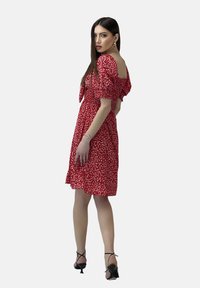 Rotes Kleid mit weißem Blumenmuster, kurzen Puffärmeln, quadratischem Ausschnitt und A-Linien-Silhouette. Kombiniert mit schwarzen Sandalen mit Riemchen.