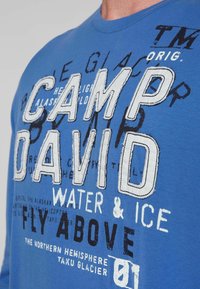 Niebieska bawełniana koszulka z dużym białym i czarnym nadrukiem oraz różnymi grafikami, w tym napisem "CAMP DAVID" oraz "WATER & ICE."