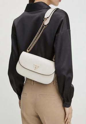 Femme portant un chemisier noir et un pantalon beige avec un sac à bandoulière blanc Guess avec des lanières en chaîne dorée dans le dos.