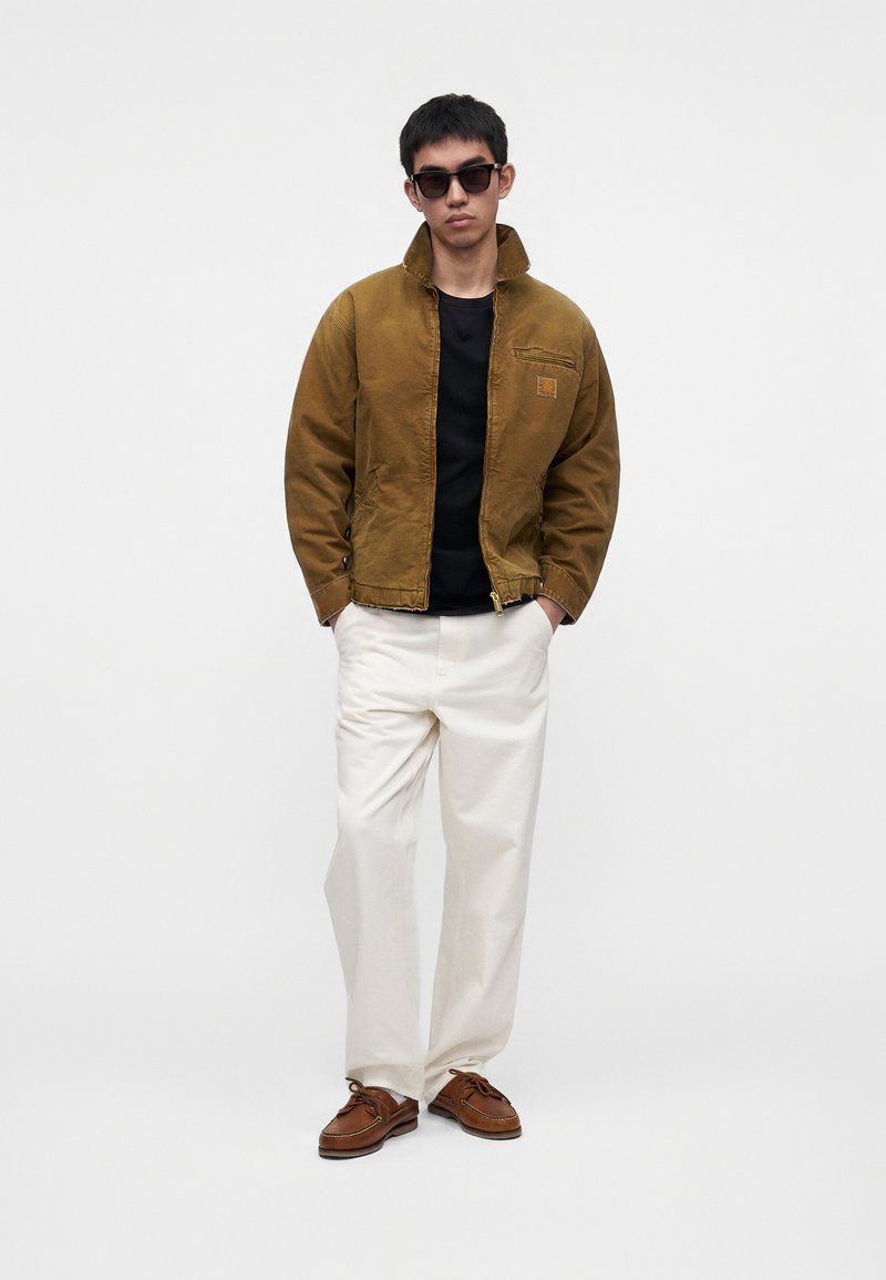Homme portant une veste marron zippée sur une chemise noire, pantalon blanc, chaussures marron et lunettes de soleil foncées, debout les mains dans les poches.