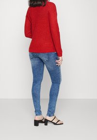 Suéter de punto rojo con textura acanalada, combinado con jeans skinny azul de tiro medio. Sandalias negras de tacón ancho completan el conjunto.