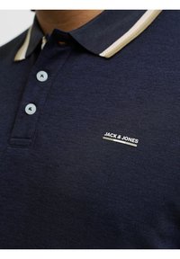 Navyblaues Poloshirt mit strukturiertem Gewebe, bestehend aus einem gestrickten Kragen in Creme, drei hellblauen Knöpfen und einem kleinen weißen Logo auf der Brust.