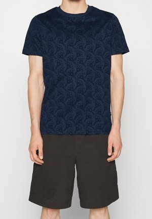 Mężczyzna ubrany w granatowy krótki T-shirt w kwiaty i czarne szorty do kolan na jednolitym tle.