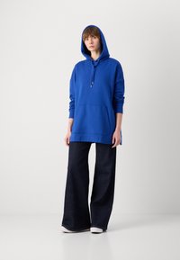 Blaue Kapuzenjacke mit Kordelzug und einer Vordertasche, kombiniert mit dunklen, weit geschnittenen Jeans. Der Stoff wirkt weich und bequem.
