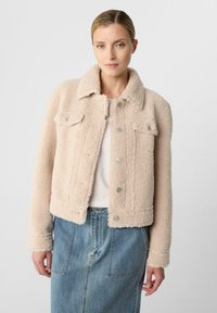 Donna che indossa una giacca sherpa beige con chiusure a bottone sopra una maglia bianca e una gonna di jeans blu, in piedi su uno sfondo neutro.
