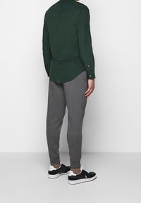 Zelená košile na knoflíky, šedé tapered tepláky a černé tenisky. Košile má hladkou texturu; tepláky jsou měkké a mírně elastické.