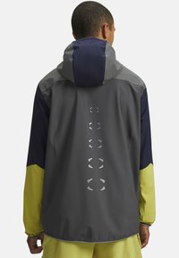 Vandafvisende jakke med hætte; har grå, navy og gule paneler; reflekterende detaljer langs ryggen; glat tekstur.