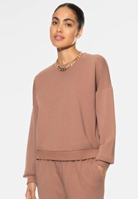 Bruine sweatshirt gemaakt van zachte stof met een relaxte pasvorm, verlaagde schouders en een geribde zoom. Gepaard met een gouden kettinghalskette.