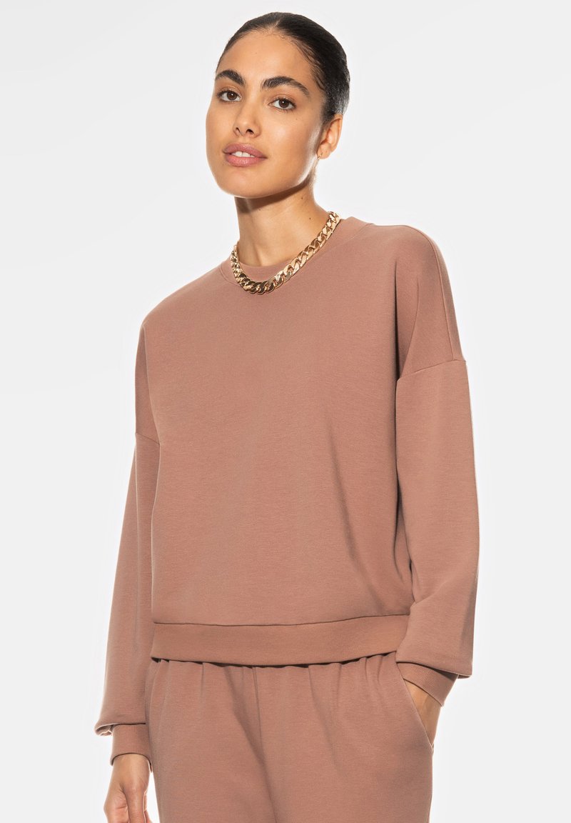 Bruine sweatshirt gemaakt van zachte stof met een relaxte pasvorm, verlaagde schouders en een geribde zoom. Gepaard met een gouden kettinghalskette.