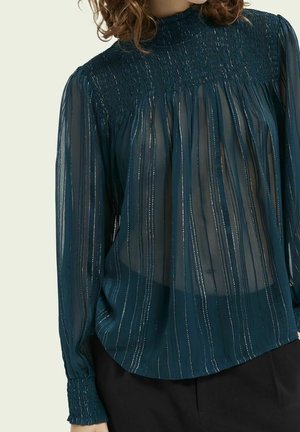 Ciemno morska przezroczysta bluzka z pionowymi metalicznymi paskami, marszczonym wysokim kołnierzem i mankietami, noszona z czarnymi spodniami na osobie o kręconych włosach.