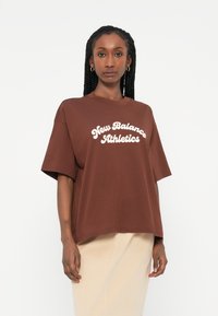 Brūns kokvilnas T-krekls ar īsām piedurknēm un apaļu dekoltē. Priekšpusē ir balti slīpā rakstā uzrakstīts "New Balance Athletics".