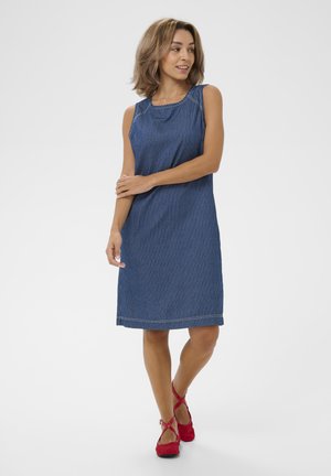 Kobieta w niebieskiej sukience w prążki bez rękawów, sięgającej do kolan, i czerwonych balerinach, stojąca ze skrzyżowanymi nogami i delikatnie złożonymi rękami.