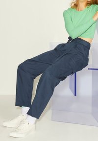 Person med högmidjade mörkblå cargobyxor, vita sneakers, grön långärmad crop top, sitter på en genomskinlig stol mot en enfärgad bakgrund.