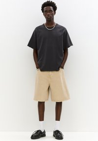 Tricou supradimensionat de culoare gri închis, cu mâneci scurte, asociat cu pantaloni scurți bej până la genunchi. Pantofi negri și șosete albe completează ținuta.