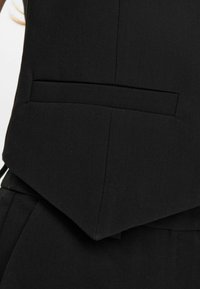 Chaleco negro a medida con un corte afilado y angular que incluye un bolsillo oculto. La tela tiene una textura suave, acentuando un diseño elegante.