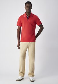 Rotes Poloshirt mit kurzen Ärmeln und navyfarbenen Kragenakzenten, kombiniert mit hellbeigen weiten Hosen und weißen Sneakers. Glatter, lässiger Stoff.
