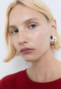 Primo piano di una persona con i capelli biondi corti, che indossa un maglione rosso e orecchini a cerchio argentati, con pelle liscia e occhi azzurri chiari.