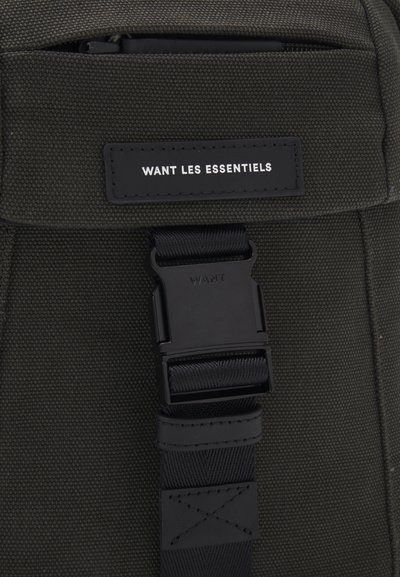 Tamnozeleni platneni ruksak s crnom plastičnom kopčom i etiketom na kojoj je ispisano "WANT LES ESSENTIELS" u bijelom. Tekstura površine.