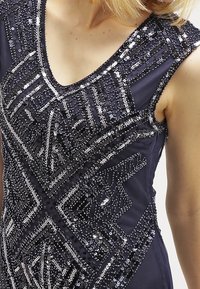 Námořnické šaty s propracovanými geometrickými vzory z flitrů. Bez rukávů, s hlubokým výstřihem do V a hladkou látkou se třpytivými prvky.