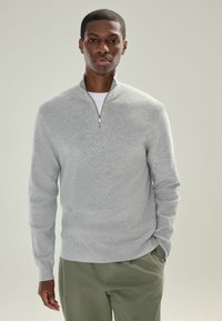 Jersey de punto gris claro con un cuello con cremallera de un cuarto, textura acanalada y mangas largas, combinado con pantalones verde oliva.