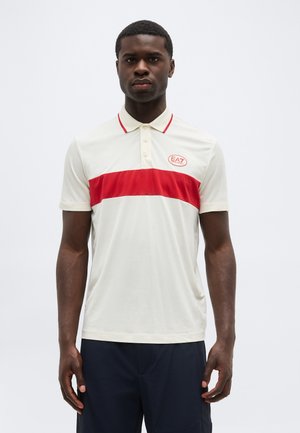 Άνδρας που φορά λευκό πόλο με κόκκινη ρίγα στο στήθος και λογότυπο EA7, σε συνδυασμό με σκούρο παντελόνι, στέκεται μπροστά σε απλό φόντο.