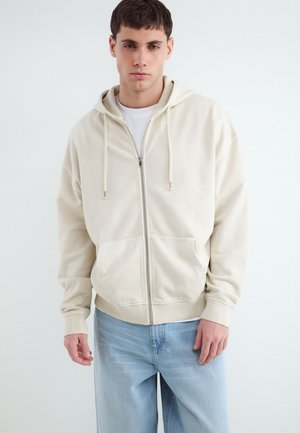 Junger Mann trägt einen beigen Kapuzenpullover mit Reißverschluss und Taschen sowie hellblaue Jeans vor weißem Hintergrund.