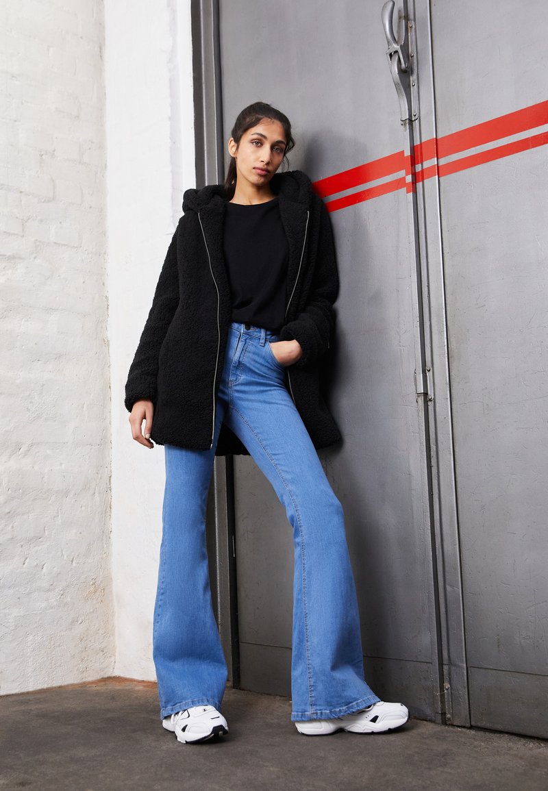 Veste noire fluffy avec capuche, associée à un jean flare bleu clair et à des sneakers blanches, sur fond gris avec des accents rouges.