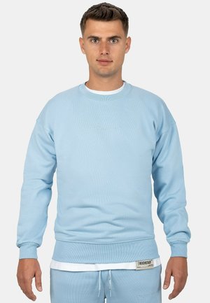 Light blue sweatshirt in soft fabric with ribbed cuffs and waistband, featuring a subtle front embroidery and layered white shirt beneath.