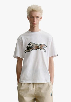 Giovane uomo con capelli chiari che indossa una maglietta bianca con un disegno di una volpe dei cartoni animati che salta in stampa mimetica e pantaloni beige con le mani in tasca.