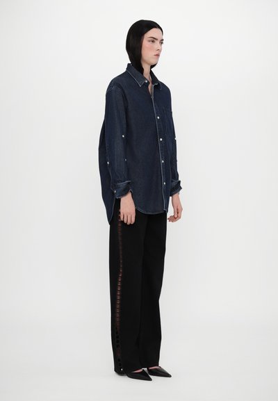 Chemise en denim oversized avec boutons sur le devant, manches longues et poche poitrine ; assortie à un pantalon noir avec des découpes circulaires sur le côté.
