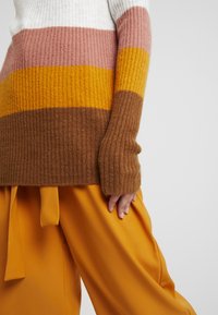Mehrfarbiger Strickpullover mit breiten horizontalen Streifen in Weiß, Pink, Gelb und Braun. Hat eine gerippte Textur und eine lockere Passform. Kombiniert mit orangenen Hosen.