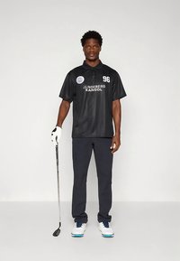 Polo desportivo preta com tecido que afasta a humidade, apresentando um logótipo e o número 96. Combinado com calças azul-marinho e sapatos de golfe brancos, segurando um taco de golfe.