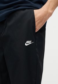 Άτομο που φοράει μαύρο παντελόνι Nike με το χέρι μέσα στην πλαϊνή τσέπη, ορατό λευκό λογότυπο Nike στον μηρό και σκούρο επάνω μέρος.