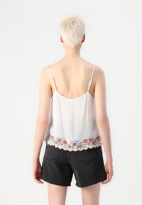Personne aux cheveux blonds courts portant un haut sans manches blanc brodé de fleurs et un short en denim noir, debout de dos.