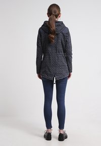 Navy-mönstrad jacka med huva, tillverkad av texturerat tyg, med en insydd midja och en längre bak. Matchad med mörka denimjeans.