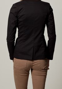 Blazer noir ajusté avec un design cintré, une fente simple au dos et un subtil détail de couture. Porté sur un pantalon beige.