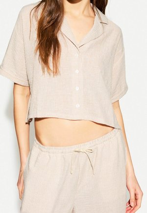 Kobieta ubrana w beżowo-białą, pionowo paskowaną, krótką koszulę na guziki oraz dopasowane spodnie ze ściągaczem, stojąca na białym tle.