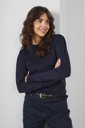Pull en maille marine à manches longues, col rond et poignets côtelés, associé à un pantalon sombre et une ceinture marron, sur un fond clair.