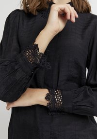 Blusa nera in tessuto testurizzato, con collo alto e maniche lunghe. Le maniche presentano sofisticate merletti alle estremità con orlo scallop.
