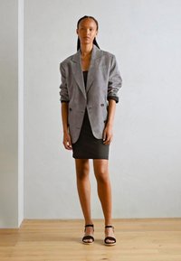 Blazer de pana gris de corte cruzado sobre un vestido negro ajustado, combinado con tacones negros de tiras. Texturas suaves, diseño a medida.